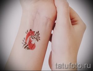 Небольшая татуировка лисы в коричнево-красных тонах на женском запястье. Рисунок выполнен в минималистичном стиле с добавлением зеленых листьев.