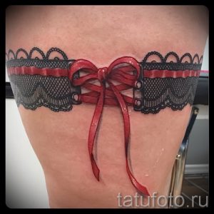 Фото тату кружевного чулка с красным бантом на бедре. Детализированный рисунок татуировки, выполненный в черно-красных тонах.