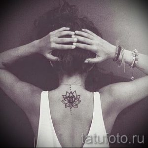 Фото тату: небольшая черная татуировка лотоса на спине, с тонкими линиями и точечным узором, расположенная между лопатками.