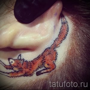 Фото тату лисы возле уха, выполненное в небольшом размере. Рисунок татуировки изображает лису в движении, с поднятой лапой.