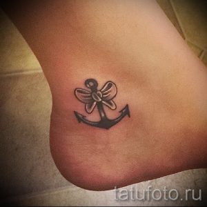 Фото тату маленького якоря, украшенного бантиком, на лодыжке. Нежный и милый рисунок татуировки, выполненный в черно-серых тонах.