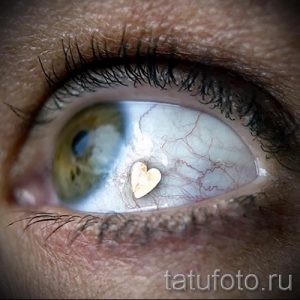 Фотография глаза с маленькой белой татуировкой в форме сердца на радужке. Крупный план, видны кровеносные сосуды.