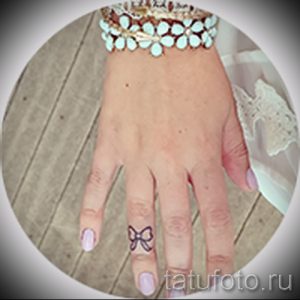 Фото тату с изображением маленького бантика на пальце девушки. Нежный и минималистичный рисунок татуировки в черном цвете.