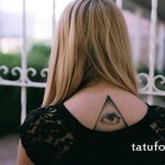 Фото минималистичной татуировки с изображением глаза в треугольнике на спине девушки. Черно-белый рисунок татуировки.