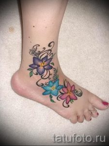 Фото татуировки на лодыжке с изображением трех ярких цветов: фиолетового, голубого и розового. Детализированный рисунок с листьями и завитушками.