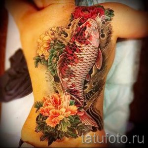 Фото тату: красочный карп кои, плывущий вверх, окруженный пышными цветами пионов на женской спине. Детализированный рисунок татуировки.