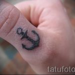 Фото маленькой татуировки на пальце: стилизованный якорь, выполненный черными чернилами. Простой и символичный рисунок.