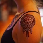 Фото татуировки мандалы на плече. Чёрно-коричневый рисунок с тонкими линиями и элементами, напоминающими капли, выполненный в изящном стиле.