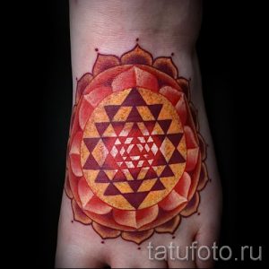 Фото татуировки Шри Янтры, выполненной в теплых оранжевых и красных тонах на лодыжке, с детальной прорисовкой геометрических фигур.
