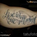 Фото тату: фраза "Live in the Moment" выполнена черным цветом на предплечье, с добавлением легких серых теней, имитирующих облака.