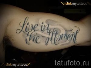 Фото тату: фраза "Live in the Moment" выполнена черным цветом на предплечье, с добавлением легких серых теней, имитирующих облака.