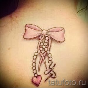 Фото тату банта с жемчужной нитью на ключице, выполненного в нежных розовых тонах. Детализированный рисунок татуировки.