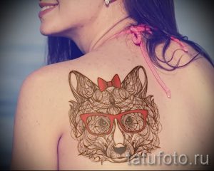 Фото татуировки лисы в очках и с бантиком на спине. Детализированный рисунок, выполненный тонкими линиями.