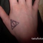 Фотография черной татуировки на ладони с изображением символа Даров Смерти из Гарри Поттера. Небольшой и лаконичный рисунок татуировки.