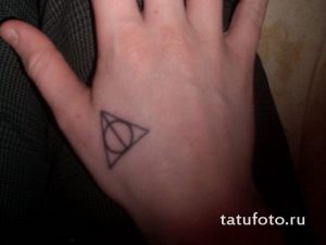 Фотография черной татуировки на ладони с изображением символа Даров Смерти из Гарри Поттера. Небольшой и лаконичный рисунок татуировки.