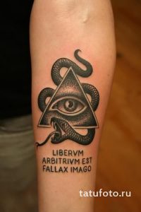 Фото тату на предплечье: Всевидящее око в треугольнике, обвитое змеей, с латинской надписью. Стильный и загадочный рисунок татуировки.