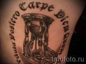 Фото тату песочных часов с латинской надписью "Carpe Diem Quam Minimum Credula Postero". Рисунок выполнен в реалистичном стиле с использованием черных и серых оттенков.