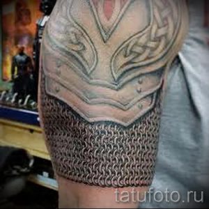 Фото тату: татуировка на плече, изображающая стилизованную броню с кельтским узором и цепным плетением, выполненная в черно-серых тонах.