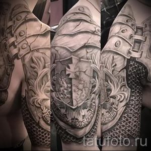 Фото тату: масштабная татуировка на плече и руке, изображающая рыцарские доспехи с гербом и цепным плетением, выполненная в черно-серых тонах.