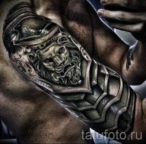 Фото тату: реалистичный рисунок льва в шлеме и броне на предплечье мужчины, выполненный в черно-серых тонах с высокой детализацией.