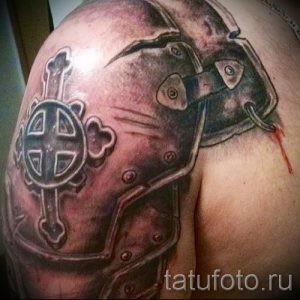 Фото тату на плече с изображением рыцарского доспеха и кельтского креста. Черно-серый рисунок с элементами крови, добавляющими драматизма.