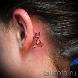 Маленькая татуировка лисы, расположенная за ухом. Изображение выполнено в ярком оранжевом цвете с использованием простых линий и форм.