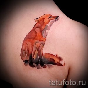 Фото тату сидящей лисы на плече, выполненное в технике акварели. Изображение сочетает в себе плавные переходы цветов и реалистичные детали.