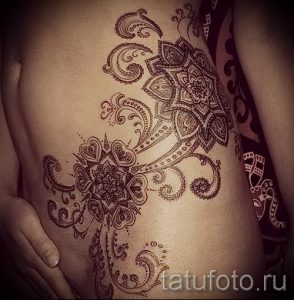 Фото татуировки в стиле мехенди на женском боку. Детализированный рисунок татуировки с использованием витиеватых узоров и цветочных мотивов в коричневых тонах.
