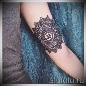 Фото татуировки мандалы с изображением символа Ом в центре, расположенной на предплечье. Рисунок выполнен в черно-белом стиле с высокой детализацией.