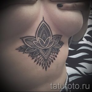 Фото тату мандалы и цветка лотоса, выполненной в технике дотворк, на женской груди. Детализированный рисунок татуировки с геометрическими узорами и тонкими линиями.