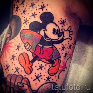 Фото тату Микки Мауса, катающегося на скейтборде, с яркими брызгами краски и звездным фоном. Рисунок татуировки выполнен в мультяшном стиле.