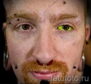 Фото мужчины с множеством пирсинга на лице, а также с татуировками и зеленым цветом в одном глазу. Демонстрация экстремальной боди-модификации.