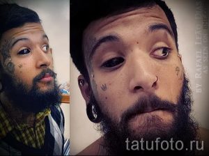 Фотография мужчины с множеством татуировок на лице, шее и теле. Татуировки выполнены в различных стилях и имеют сложный узор.