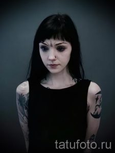 Фотография девушки с темными волосами и множеством татуировок на теле, включая рисунки на руках и лице. Пирсинг на носу добавляет образу загадочности.