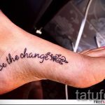 Фото тату: черная надпись "Be the change" с небольшими растительными элементами на женской лодыжке.