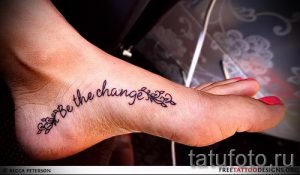 Фото тату: черная надпись "Be the change" с небольшими растительными элементами на женской лодыжке.
