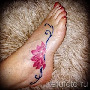 Фото тату розового лотоса на стопе. Изображение цветка с изящными лепестками и синими волнистыми линиями. Минималистичный рисунок татуировки.