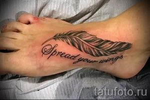 Фото тату на стопе: черное перо с надписью 'Spread your wings'. Мотивирующий рисунок татуировки в стиле каллиграфия.