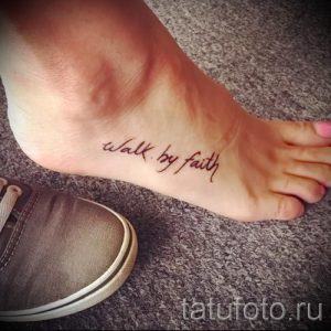 Фото тату надписи 'walk by faith' на стопе. Коричневый цвет, каллиграфический шрифт, религиозный стиль. Подходит для верующих людей.
