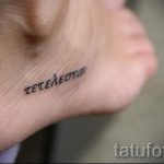 Фото тату надписи на греческом языке на внутренней стороне стопы. Черный контур, тонкие линии, минималистичный стиль. Небольшой размер.