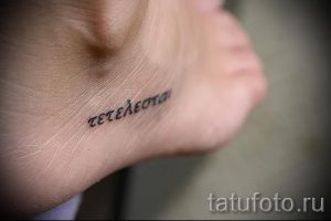 Фото тату надписи на греческом языке на внутренней стороне стопы. Черный контур, тонкие линии, минималистичный стиль. Небольшой размер.