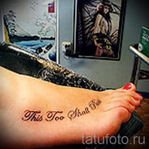 Фото тату: Татуировка с надписью "This Too Shall Pass" на стопе, выполненная в каллиграфическом стиле. Вдохновляющий и позитивный рисунок татуировки.