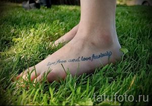 Фото тату надписи 'Live free and love fearlessly' на женской стопе. Надпись выполнена голубым цветом, на фоне зеленой травы.
