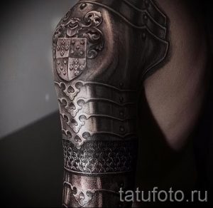 Фото тату, изображающего герб на плече. Герб выполнен в черно-серых тонах с детальной проработкой элементов.