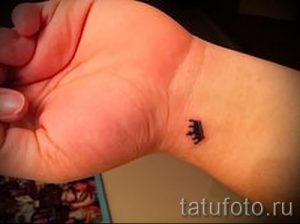 Фотография женского запястья с маленькой татуировкой в виде черной короны, выполненной в минималистичном стиле.