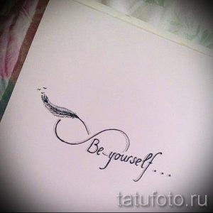 Фото минималистичной татуировки в виде символа бесконечности, внутри которого расположено перо и надпись 'Be yourself'. Черно-белый рисунок, символизирующий свободу и индивидуальность.