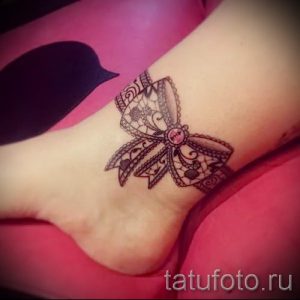 Фото татуировки в виде кружевного банта с небольшим камнем на лодыжке. Нежный и романтичный рисунок.