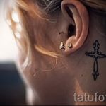 Фото тату креста с декоративными завитушками, расположенного за ухом. Изящный и женственный рисунок татуировки в черном цвете.