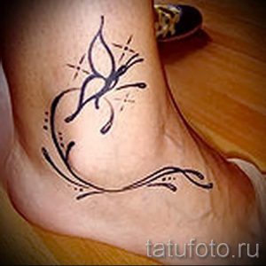 Фото татуировки с изображением черной бабочки и декоративных завитков на лодыжке, выполненной в графическом стиле.