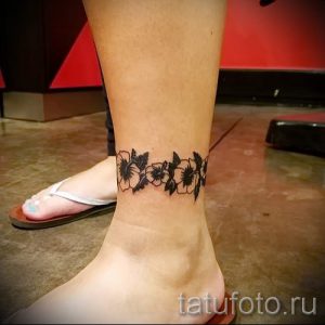Фото тату цветочного браслета на лодыжке. Черно-белый рисунок татуировки с детальной проработкой лепестков и листьев.